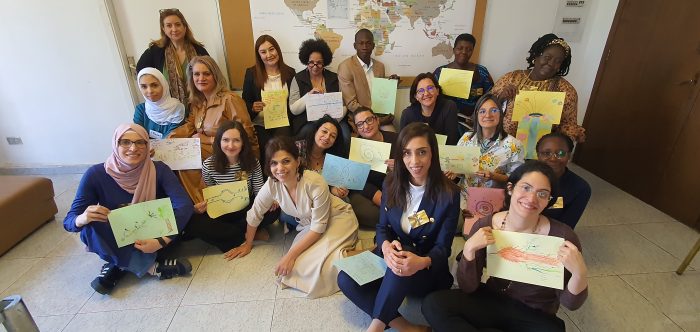 A group of people sitting down on the floor holding drawings in different colours, with a map of the world behind them