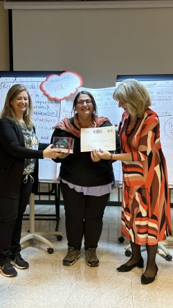 Three women standing together and smiling, flipchart paper and a sign "Welcome IALLA" is behind them, the one in the middle is holding a certificate and a small sculpture with a tree