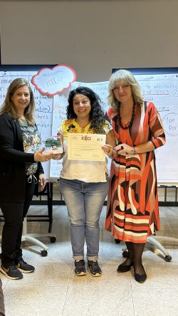 Three women standing together, flipchart paper and a sign "Welcome IALLA" behind them, the one in the middle is holding a certificate and a small sculpture of a tree