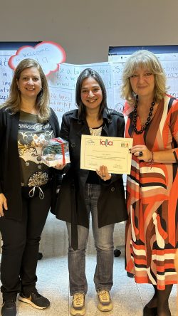 Three women standing together in front of flipchart paper, they're smiling, the one in the middle is holding a certificate