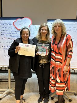 Three women standing together and smiling, flipchart paper and sign "Welcome IALLA" behind them, the one on the left is holding a certificate