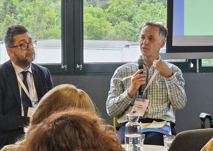 a man speaking and holding a microphone, other man listening